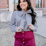 Woman with dark hair standing in a front profile position wearing the Sadie Top in a lifestyle scene. The focal point of this image is the black gingham pattern and bow that's tied on front of top. The model paired this top with a maroon skirt. 