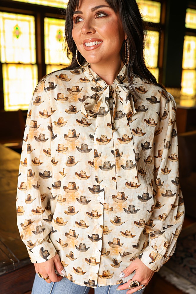 Woman with dark hair wearing a long-sleeve button-down with multi-colored cowboy hats and tying feature in front of top. Model paired this top with light-wash denim jeans. 