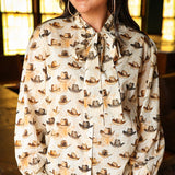 Woman with dark hair wearing a long-sleeve button-down with multi-colored cowboy hats and tying feature in front of top. Model paired this top with light-wash denim jeans. 
