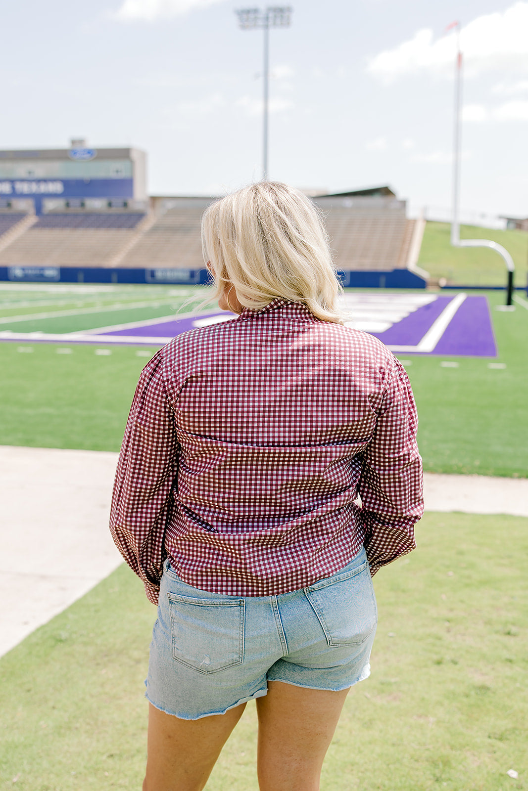 Woman with blonde hair standing in a back profile position wearing the Sadie Top in a lifestyle scene. The focal point of this image is the maroon gingham pattern on back of long-sleeve top. The model paired this top with light-wash denim shorts. 