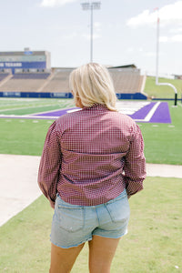 Woman with blonde hair standing in a back profile position wearing the Sadie Top in a lifestyle scene. The focal point of this image is the maroon gingham pattern on back of long-sleeve top. The model paired this top with light-wash denim shorts. 