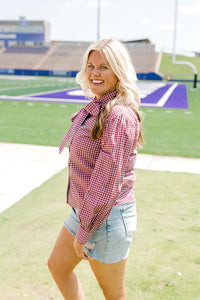 Woman with blonde hair standing in a side profile position wearing the Sadie Top in a lifestyle scene. The focal point of this image is the maroon gingham pattern along shirt, cuffed sleeve, and bow tied in front. The model paired this top with light-wash denim shorts. 