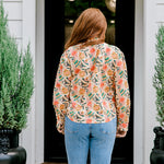 Woman with red hair wearing a long-sleeve top with multi-color ornament graphics & a bow-tied in the front. Model paired this top with light-wash denim jeans. 