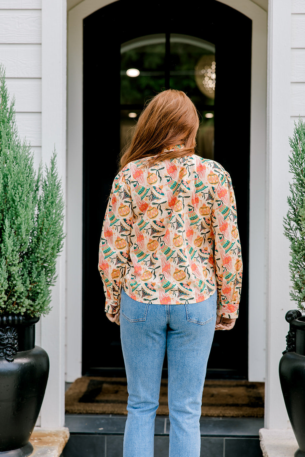 Woman with red hair wearing a long-sleeve top with multi-color ornament graphics & a bow-tied in the front. Model paired this top with light-wash denim jeans. 