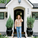 Woman with red hair wearing a long-sleeve top with multi-color ornament graphics & a bow-tied in the front. Model paired this top with light-wash denim jeans. 