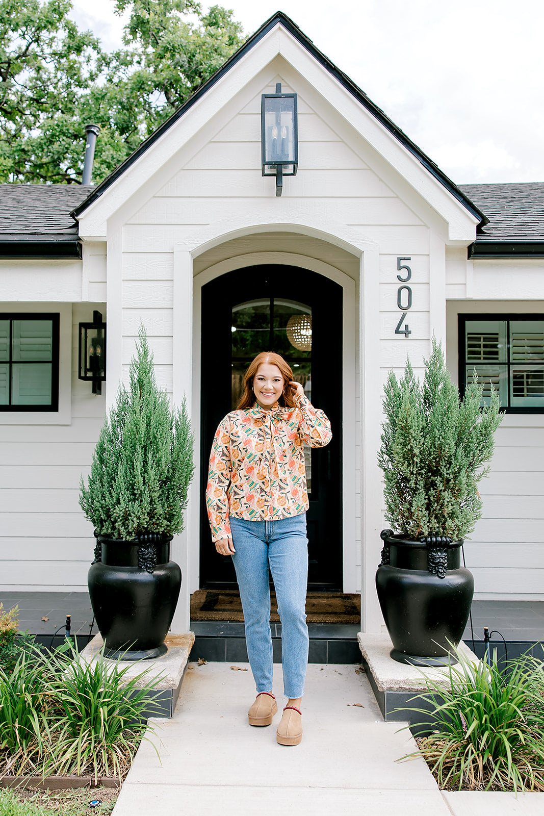 Woman with red hair wearing a long-sleeve top with multi-color ornament graphics & a bow-tied in the front. Model paired this top with light-wash denim jeans. 