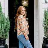 Woman with red hair wearing a long-sleeve top with multi-color ornament graphics & a bow-tied in the front. Model paired this top with light-wash denim jeans. 