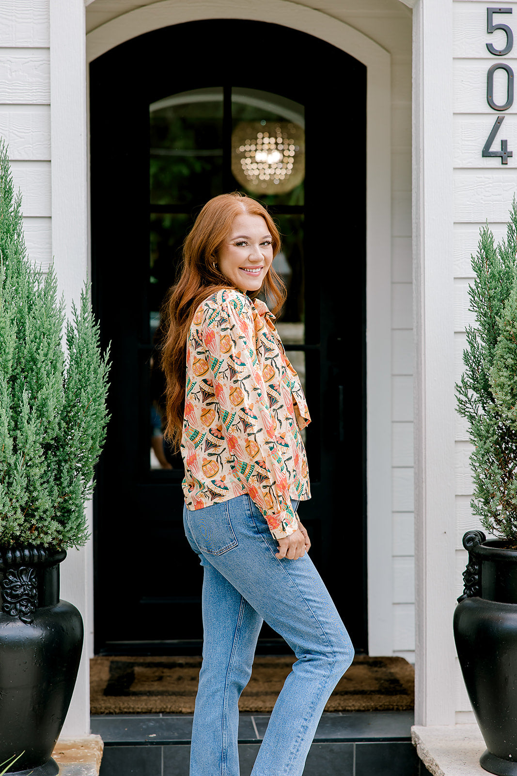 Woman with red hair wearing a long-sleeve top with multi-color ornament graphics & a bow-tied in the front. Model paired this top with light-wash denim jeans. 