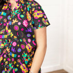 Woman with dark hair in a front facing position with a deep focal length on the front of top. The focal point of this image is the graphics on the front of the top with slight emphasis on the sleeves and ruffled collar.