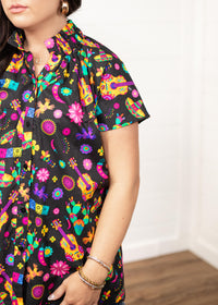 Woman with dark hair in a front facing position with a deep focal length on the front of top. The focal point of this image is the graphics on the front of the top with slight emphasis on the sleeves and ruffled collar.