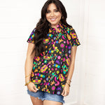 Woman with dark hair standing in a front profile position wearing the Sarah Top against a white background. The focal point of this image is the graphics on front of top with slight emphasis on button down portion. Model paired this top with dark-wash denim shorts.