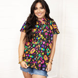 Woman with dark hair standing in a front profile position wearing the Sarah Top against a white background. The focal point of this image is the graphics on front of top with slight emphasis on button down portion. Model paired this top with dark-wash denim shorts.