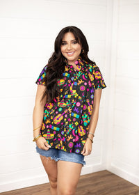 Woman with dark hair standing in a front profile position wearing the Sarah Top against a white background. The focal point of this image is the graphics on front of top with slight emphasis on button down portion. Model paired this top with dark-wash denim shorts.
