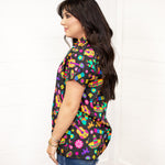 Model with dark hair wearing the Sarah Top standing in a side profile position against a white background. The focal point of this image is the side graphics on the tops, with slight emphasis on sleeves. The model paired this top with dark-wash denim shorts.