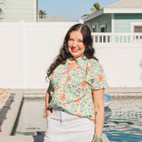 Woman with dark hair standing in a front profile position wearing the Sarah Top in a lifestyle scene. The focal point of this image is to the see the top in its entirety as well as give it a "real time" look. Model is wearing a white denim skirt to pair with top.