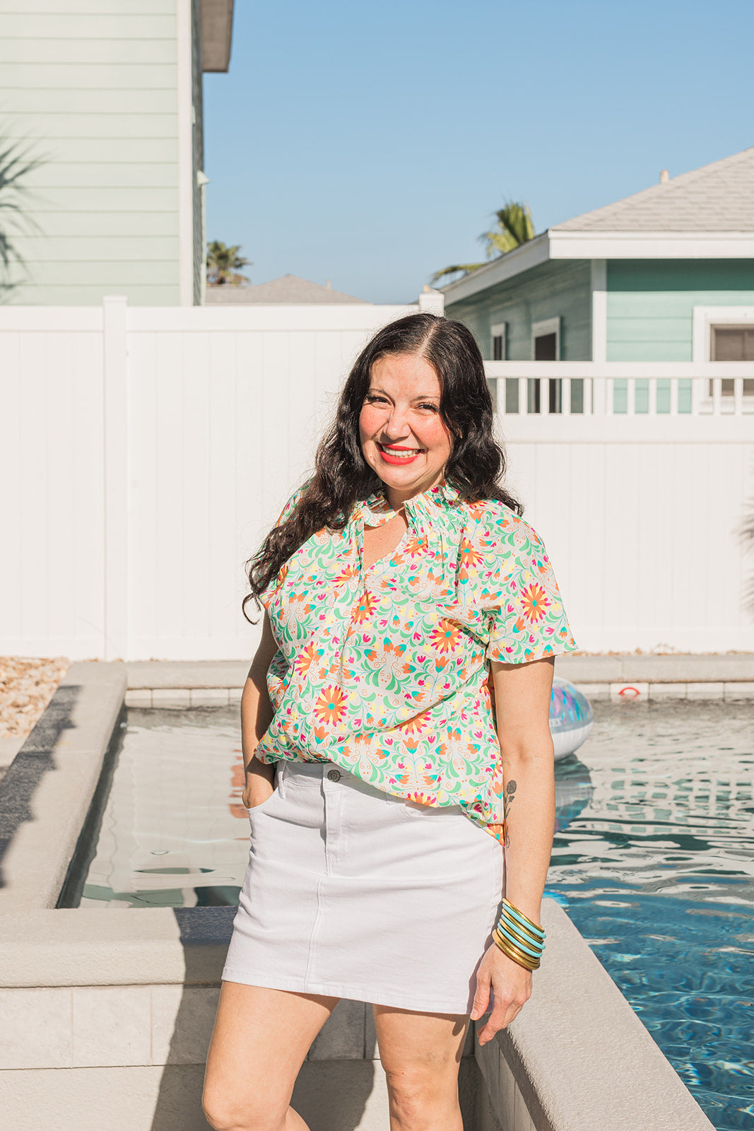 Woman with dark hair standing in a front profile position wearing the Sarah Top in a lifestyle scene. The focal point of this image is to the see the top in its entirety as well as give it a "real time" look. Model is wearing a white denim skirt to pair with top.