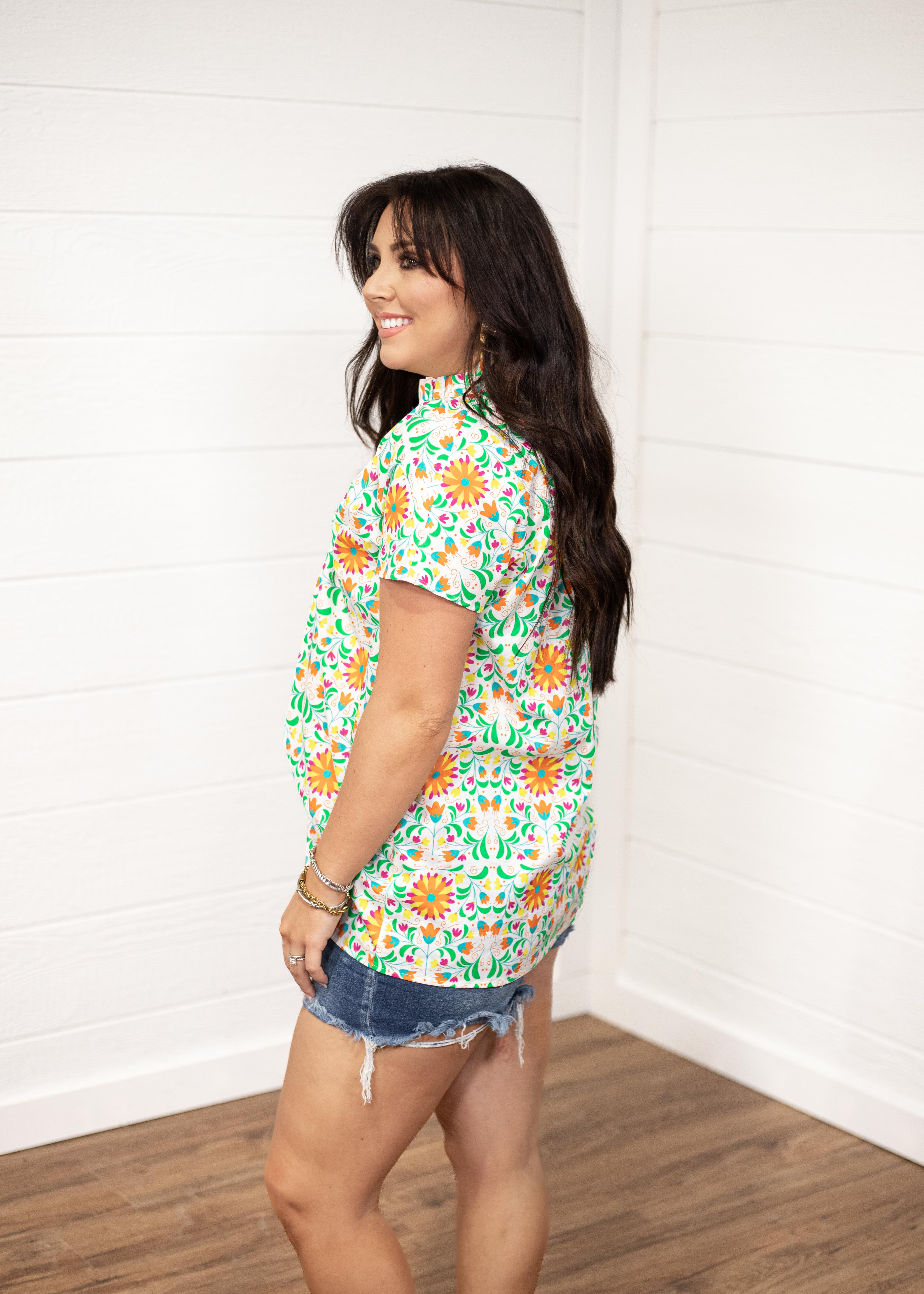 Woman with dark hair standing in a side profile position wearing the Sarah Top against a white background. The focal point of this image is the side graphics of the top as well as seeing length of top and sleeves. The model paired this top with dark-wash denim shorts.