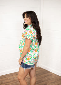 Woman with dark hair standing in a side profile position wearing the Sarah Top against a white background. The focal point of this image is the side graphics of the top as well as seeing length of top and sleeves. The model paired this top with dark-wash denim shorts.
