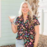 Woman with blonde hair standing in a front profile position in a lifestyle scene wearing the Sarah Top. The background of this image is backyard style gathering with emphasis on bright colors. The model paired this top with light-wash denim shorts.