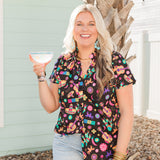 Woman with blonde hair standing in a front profile position in a lifestyle scene wearing the Sarah Top. The background of this image is backyard style gathering with emphasis on bright colors. The model paired this top with light-wash denim shorts.