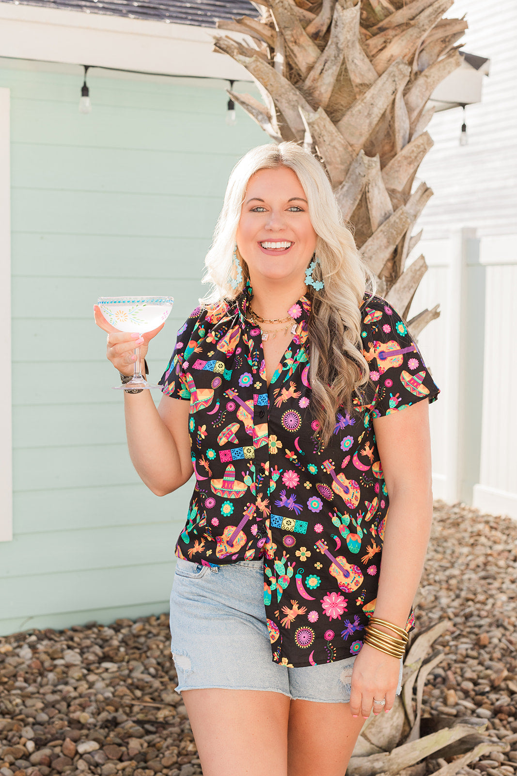 Woman with blonde hair standing in a front profile position in a lifestyle scene wearing the Sarah Top. The background of this image is backyard style gathering with emphasis on bright colors. The model paired this top with light-wash denim shorts.