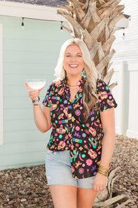 Woman with blonde hair standing in a front profile position in a lifestyle scene wearing the Sarah Top. The background of this image is backyard style gathering with emphasis on bright colors. The model paired this top with light-wash denim shorts.