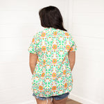 Woman with dark hair standing in a back profile position wearing the Sarah Top against a white background. The focal point of the image is the color floral designs on the back of the top. The model paired this top with dark-wash denim shorts.