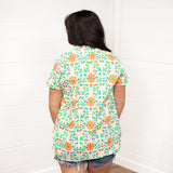 Woman with dark hair standing in a back profile position wearing the Sarah Top against a white background. The focal point of the image is the color floral designs on the back of the top. The model paired this top with dark-wash denim shorts.
