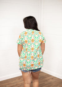Woman with dark hair standing in a back profile position wearing the Sarah Top against a white background. The focal point of the image is the color floral designs on the back of the top. The model paired this top with dark-wash denim shorts.