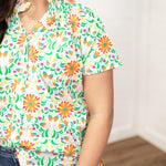 Woman with dark hair standing in a front profile position wearing the Sarah Top against a white background. The focal point of this image is to get a better look at the detail of the graphic on the top as well as the ruffles in the collar. The model paired this top with dark-wash denim shorts.