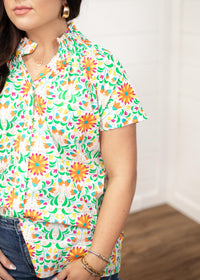 Woman with dark hair standing in a front profile position wearing the Sarah Top against a white background. The focal point of this image is to get a better look at the detail of the graphic on the top as well as the ruffles in the collar. The model paired this top with dark-wash denim shorts.