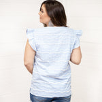 Woman with dark hair standing in a back profile position wearing the Scarlett Top against a white background. The focal point of this image is the fluffed, 3-D designs on top (blue and white). The model paired this top with dark-wash denim shorts.