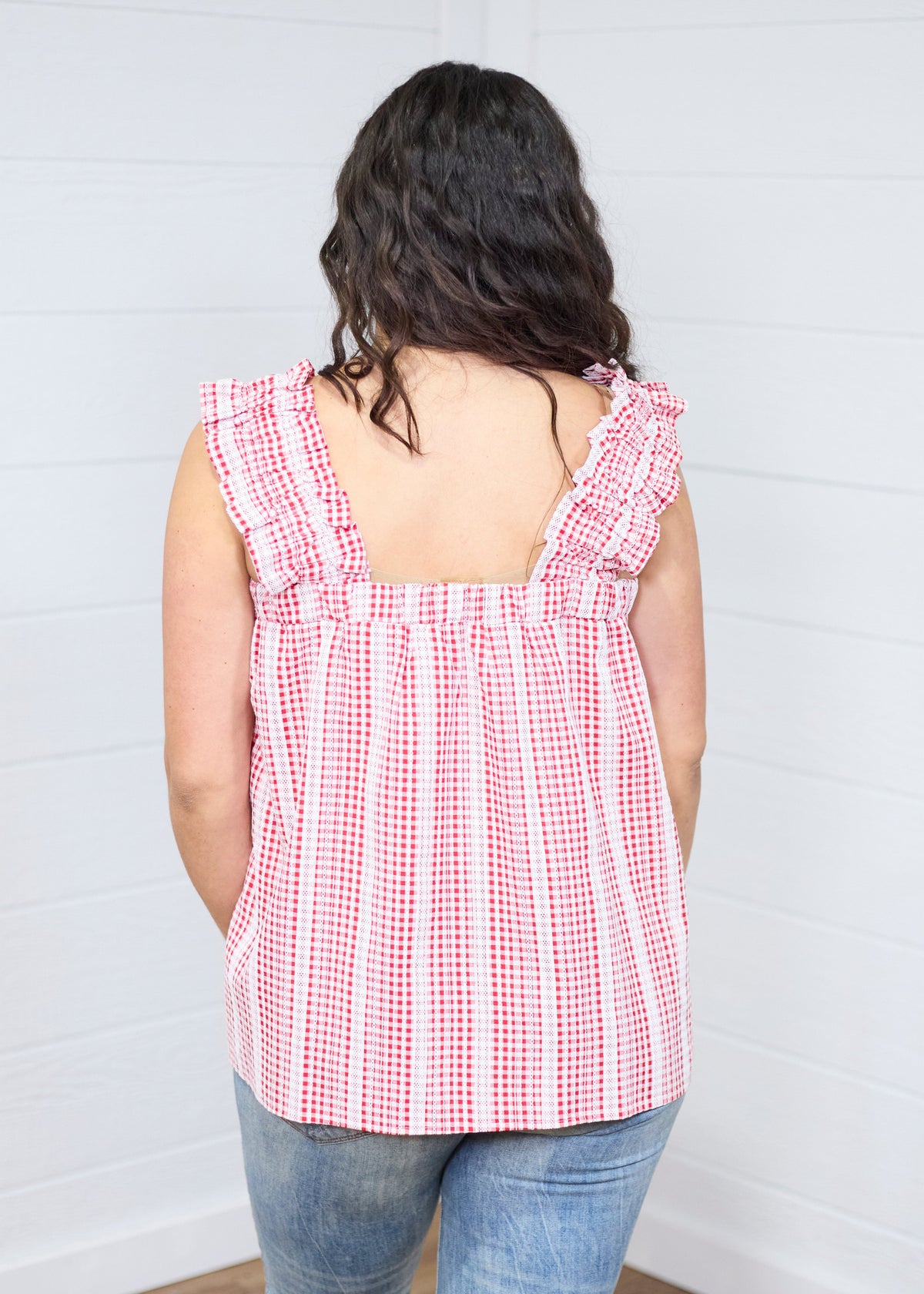 Woman with dark hair standing in a back profile position wearing the Sophia Top against a white background. The focal point for this image is the ruffled straps and gingham graphics. The model paired this top with light-wash denim jeans. 