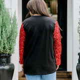 Woman with dark hair wearing long-sleeve top with embroidered cowboy Santa on front and red sparkle sleeves. Model paired this top with light-wash denim jeans. 