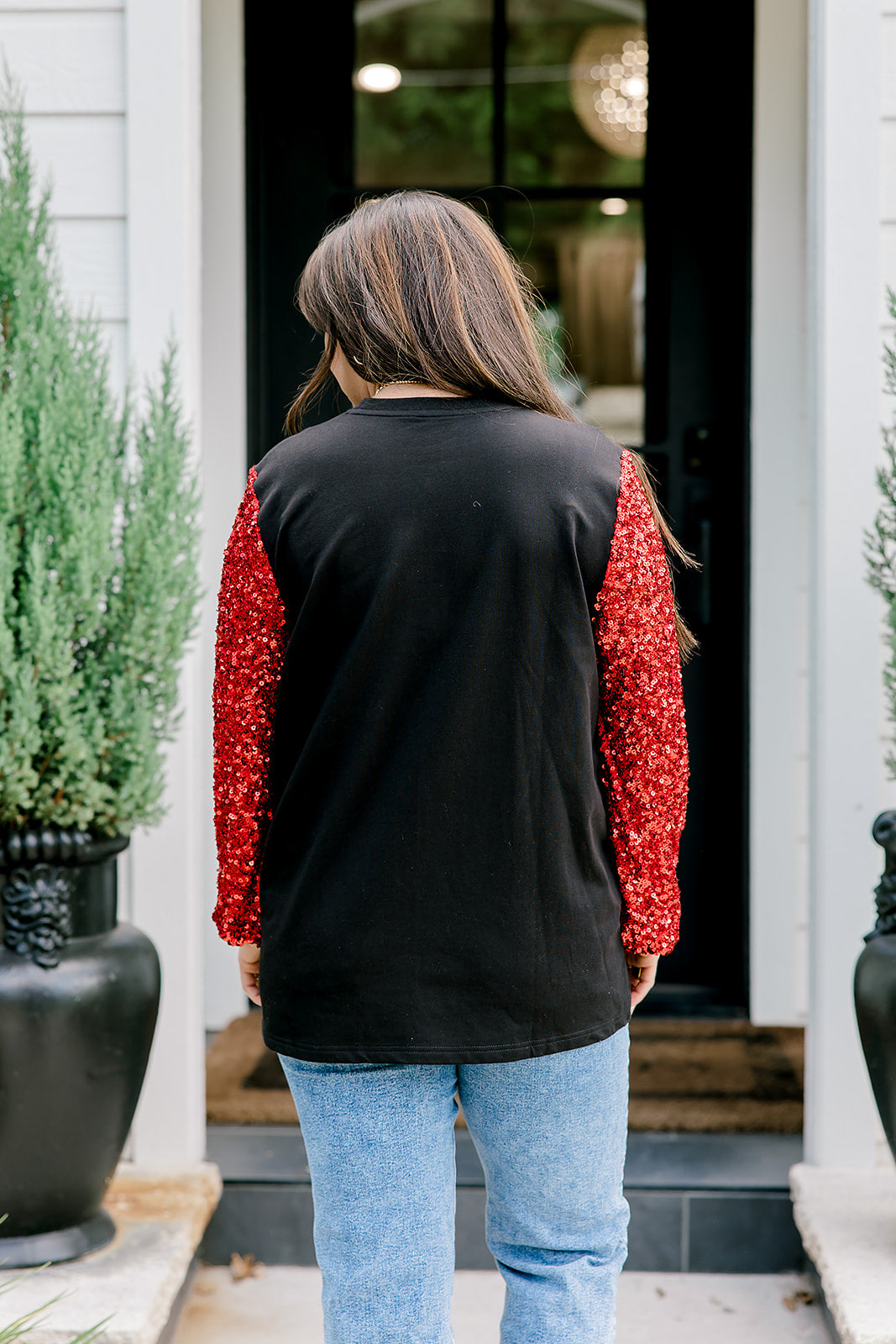 Woman with dark hair wearing long-sleeve top with embroidered cowboy Santa on front and red sparkle sleeves. Model paired this top with light-wash denim jeans. 