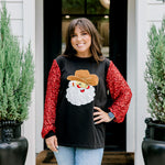 Woman with dark hair wearing long-sleeve top with embroidered cowboy Santa on front and red sparkle sleeves. Model paired this top with light-wash denim jeans. 