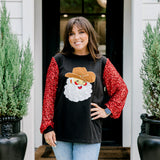 Woman with dark hair wearing long-sleeve top with embroidered cowboy Santa on front and red sparkle sleeves. Model paired this top with light-wash denim jeans. 