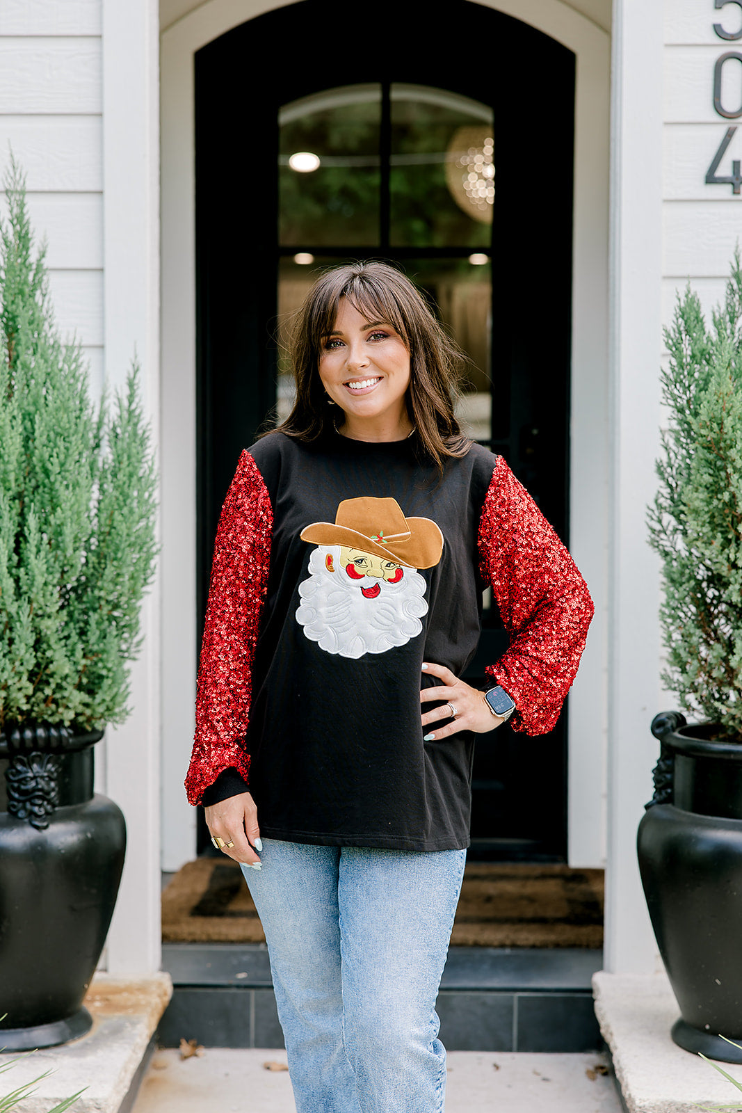 Woman with dark hair wearing long-sleeve top with embroidered cowboy Santa on front and red sparkle sleeves. Model paired this top with light-wash denim jeans. 