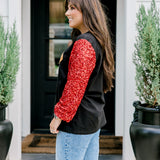Woman with dark hair wearing long-sleeve top with embroidered cowboy Santa on front and red sparkle sleeves. Model paired this top with light-wash denim jeans. 