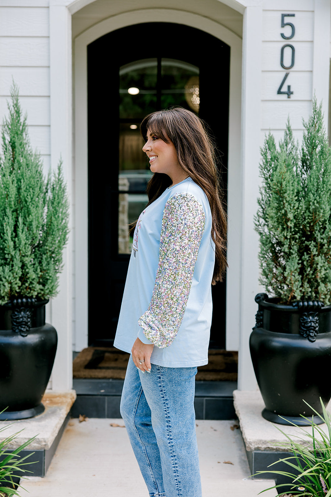 Woman with dark hair wearing a baby blue sweatshirt with iridescent sparkle sleeves; front of top has holiday truck embroidered on it. Model paired this top with light-wash denim jeans. 