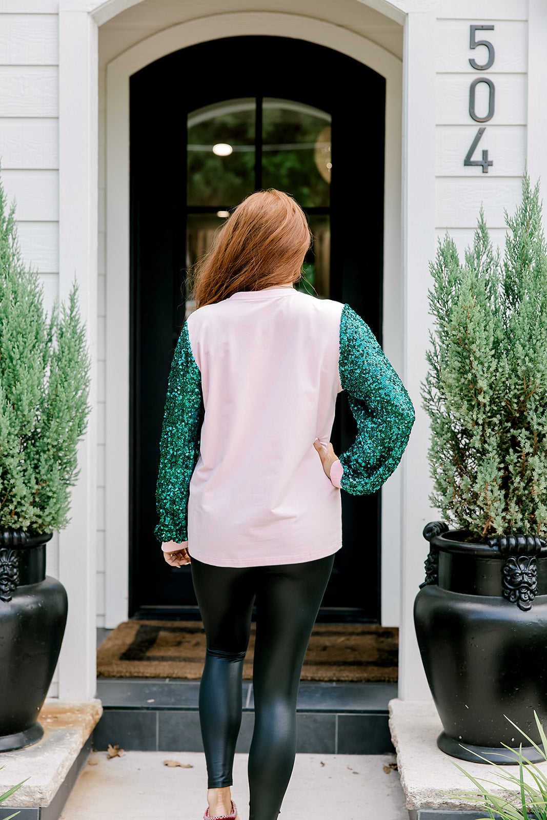 Woman with red hair wearing a sweater with green sparkle sleeves, and multi-color tree embroidery on front with words "Merry & Bright" on front. Model paired this top with faux leather leggings. 