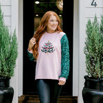 Woman with red hair wearing a sweater with green sparkle sleeves, and multi-color tree embroidery on front with words "Merry & Bright" on front. Model paired this top with faux leather leggings. 