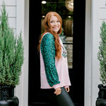 Woman with red hair wearing a sweater with green sparkle sleeves, and multi-color tree embroidery on front with words "Merry & Bright" on front. Model paired this top with faux leather leggings. 
