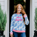 Woman with red hair wearing a blue floral sweatshirt with pink accents and "Merry Christmas" embroidered on front. Model paired this top with denim jeans. 