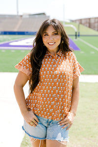 Woman with dark hair facing forwards wearing a short-sleeve, burnt orange top with flower graphics and light-wash denim shorts. 