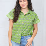 Woman with dark hair standing in a front profile position wearing the Stevie Top against a white background. The focal point of this image is the buttons down the front of the top as well as graphics and collared neckline. The model paired this top with light-wash denim jeans.
