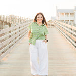 Woman with red hair standing in a front profile position wearing the Stevie Top in a lifestyle scene. The focal point of this image is the top in its entirety. The model paired this top with white linen patns.