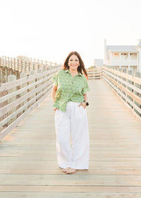 Woman with red hair standing in a front profile position wearing the Stevie Top in a lifestyle scene. The focal point of this image is the top in its entirety. The model paired this top with white linen patns.