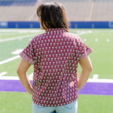 Woman with dark hair standing in a back profile position wearing the Stevie Top in a lifestyle scene. The focal point of this image is the striped ruffle sleeves, and flower graphics on the back of top. The model paired this top with light-wash denim shorts. 