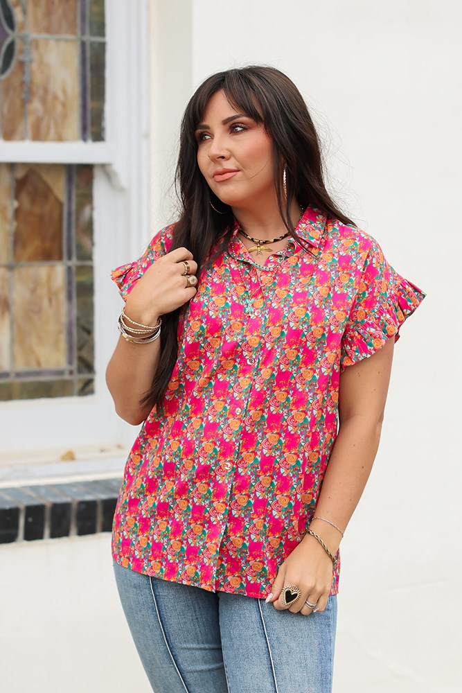 Woman with dark hair standing in a front profile position wearing the Stevie Top against a white background. The focal point of this image is the pink and orange floral, buttons down the front, as well as the low collar and ruffled sleeves. The model paired this top with dark-wash denim jeans. 